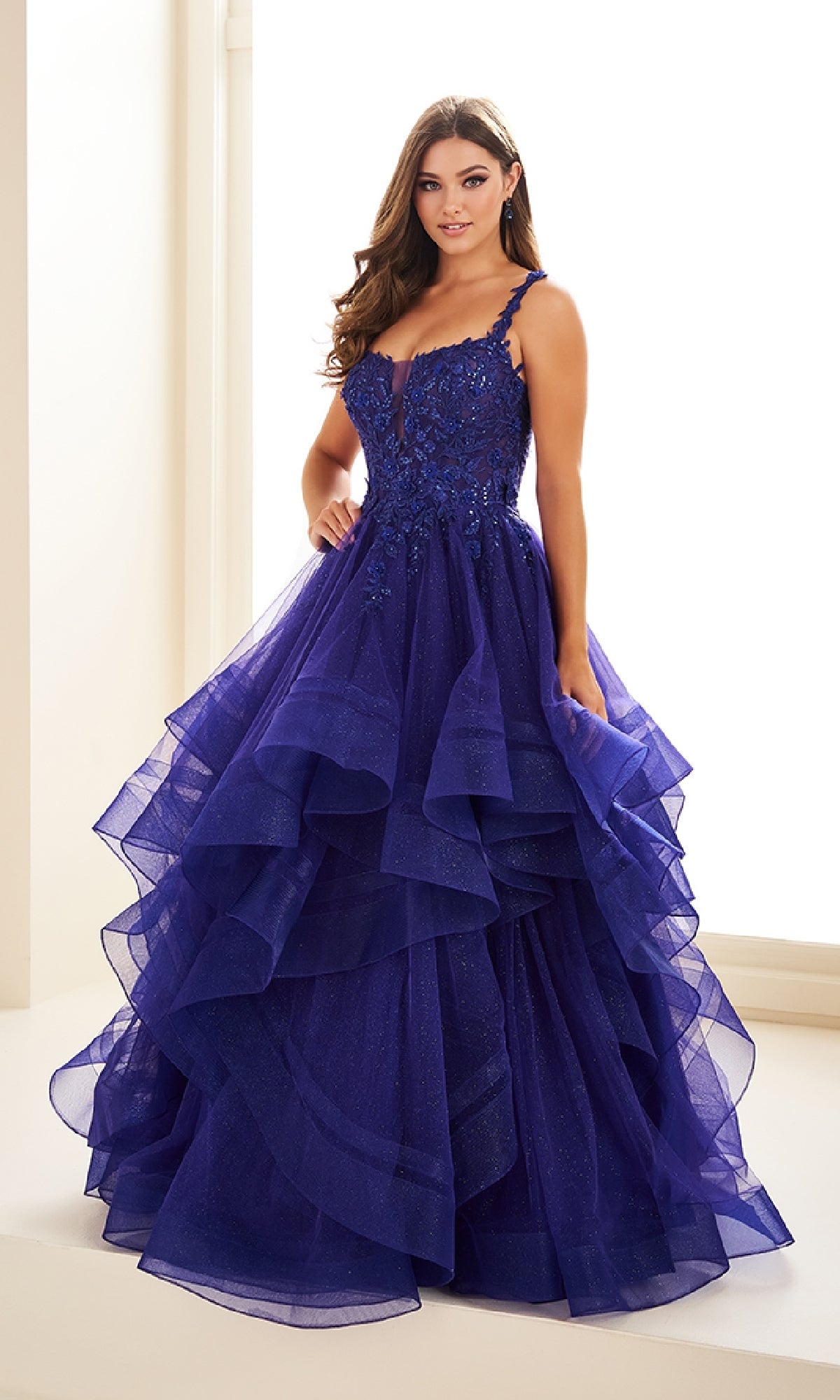 A woman wears the Ellie Wilde EW36035, a long glitter prom ball gown in royal blue, featuring a tiered A-line silhouette, floral embellishments, sequin-lace bodice, thin straps, and floor-length design.